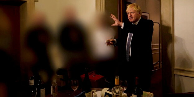 Johnson gestikuliert mit einem Glas in der Hand, unscharfe Partygäste, Alkoholflaschen auf dem Tisch