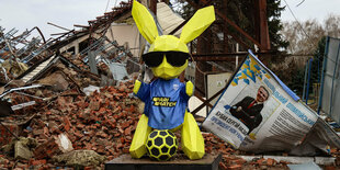 Ein knallgelbes Hasen-Maskottchen mit blauer Weste vor dem kriegszersörten Stadion in Tschernihiw