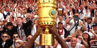 Der DFB-Pokal, ein goldener Pott, vor einer Menge an rot-weiß gekleideten Fußballfans