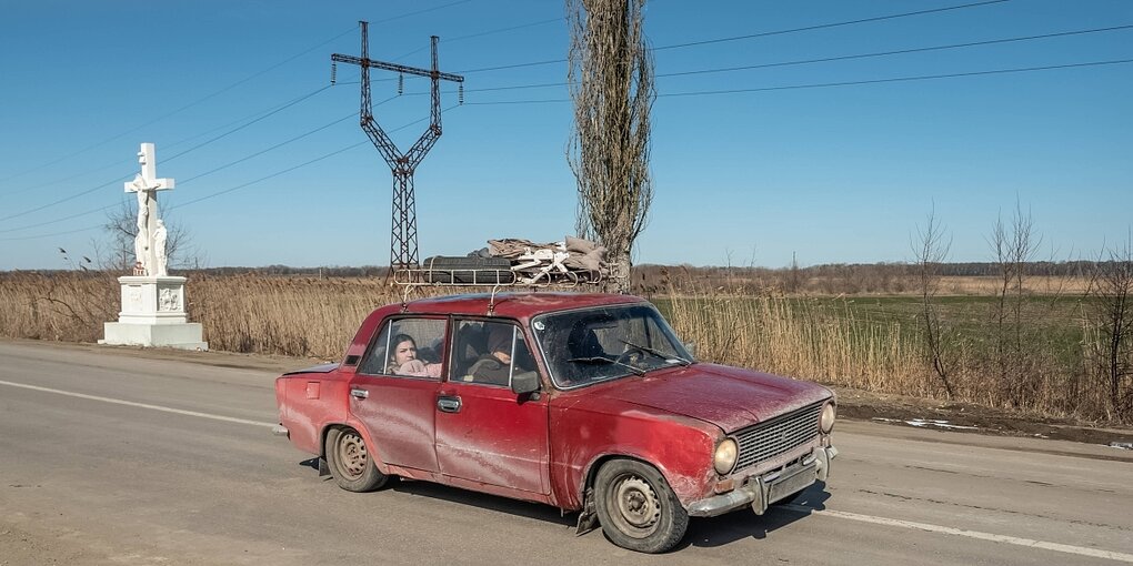 Ein rotes Auto fährt durch eine karge Landschaft. Am Wegrand steht ein weißes Kreuz