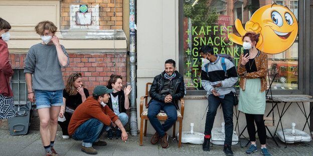 Acht Menschen stehen und sitzen vor dem "Kiez-Döner", die meisten lachen und tragen Masken. Auf der Fenster-Scheibe des "Kiez-Döner" klebt ein Emoji, der Zeigefinger und Daumen aneinander drückt