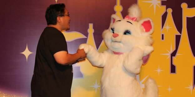 Ein Mann schüttelt einer Person im Kostüm der Aristocats-Figur Marie im Hotel von Disneyland Hong Kong die Hand. Marie, das weiße Kätzchen, trägt eine pinke Schleife und fasst sich entzückt ans Kinn