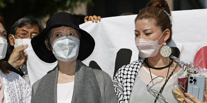 Frau mit Hut und Maske neben Frau ohne Hut und mit Maske