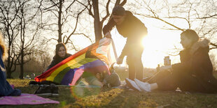 Vier Menschen sitzen auf einer Wiese. Hinter ihnen geht die Sonne unter, die Bäume haben noch keine Blätter. Paulino steht auf der Wiese und hält eine Progress-Flag in den Händen.