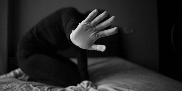 Eine junge Frau kniet auf einem Bett und hält eine Hand mit Handschuh abwehrend in die Kamera
