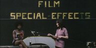 Eine Schwarze und eine weiße Frau beim Filmdreh, dahinter der Schriftzug "Film / Special-Effects": Eine Frau sitzt auf einer Bank und liest aus Valeri Solanas "SCUM" vor, daneben steht die Filmemacherin Ingelmo Engström und hält ein Mikrofon an einer Stan