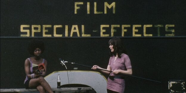 Eine Schwarze und eine weiße Frau beim Filmdreh, dahinter der Schriftzug "Film / Special-Effects": Eine Frau sitzt auf einer Bank und liest aus Valeri Solanas "SCUM" vor, daneben steht die Filmemacherin Ingelmo Engström und hält ein Mikrofon an einer Stange in Richtung der Lesenden