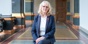 Frau sitzt im Gropius-Bau in blauem Sakko, mit blonden, mittellangen Haaren und Brille. Um sie herum sieht man Marmorböden und -wände in unterschiedlichen Farben.