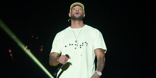 Der Rapper Booba steht auf einer dunklen Bühne und hält ein Mikrophon in der Hand