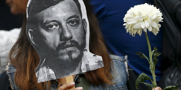 Eine Person hält eine Maske mit dem aufgemalten Gesicht von Ruben Espinosa und eine weiße Rose