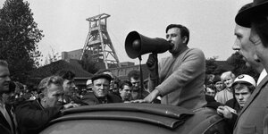 Ein historisches Foto von einer Streikszene. Arbeiter stützen sich auf ein Auto, aus dem ein Mann mit Megaphon spricht.