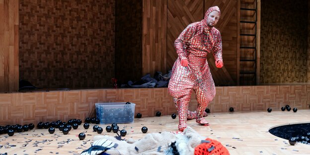 Ein Schauspieler steht in einem schuppigen Kostüm mit Salamanderschwanz auf einer Bühne voller Bälle und Lumpen