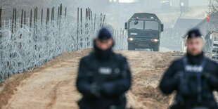 Im Vordergrund gehen zwei Grenzbeamte an einem Stacheldrahtzaun entlang. Im Hintergrund folgt ihnen ein Militärfahrzeug