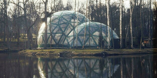 Am Ufer eines Gewässers, zwischen kahlen Bäumen, sind zwei aufblasbare, transparente Kuppeln aufgebaut