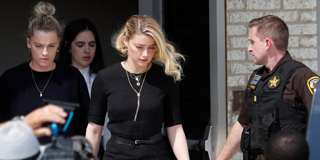 Eine blonde Frau in schwarzem Kleid verlässt ein Gebäude. Hinter ihr zwei weitere Frauen und ein Polizist