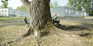Ein älterer Mann und ein Junge sitzen unter einem Baum, dahinter Baracken