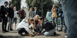 Drei Jugendliche putzen einen Stolperstein, der Rest der Klasse schaut zu