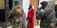 Eine Frau mit Maske steht vor einem Haus. Vor Ihr stehen Soldaten.