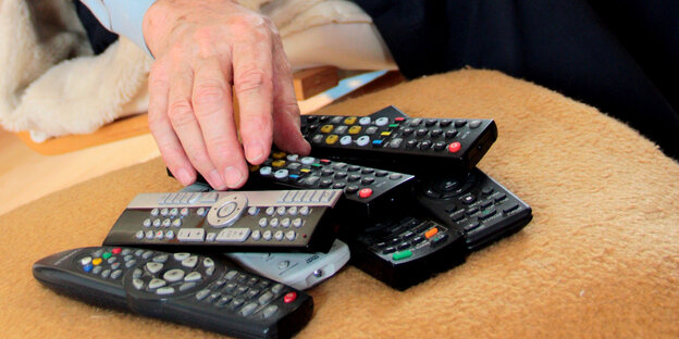 Eine Hand greift nach einem Stapel aus sieben schwarzen und grauen Fernbedienungen für den Fernseher.