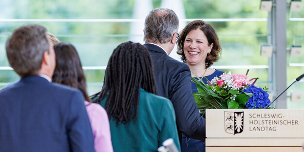 An einem Pult steht eine Frau mit Blumen in der Hand und lacht. Vor ihr steht eine Reihe von Menschen an, um ihr zu gratulieren