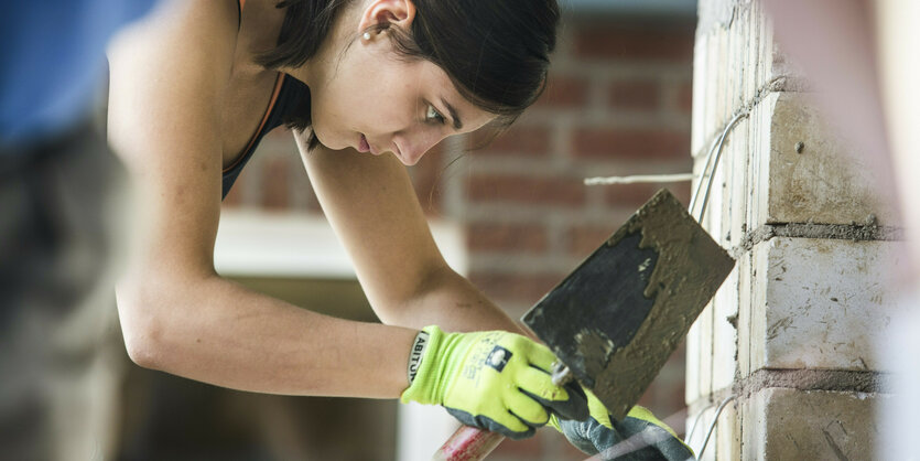 Eine Frau mit dunklen Haaren beugt sich zu einer Mauer hin und schaut konzentriert auf ihre Arbeit. In der behandschuhten Hand hält sie eine Glättkelle, mit der sie die Mauer bearbeitet.