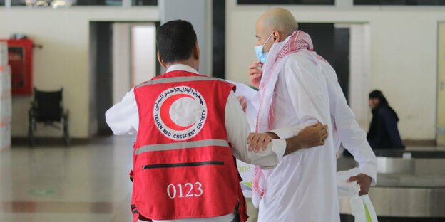 Ein Mann in einer Weste des Roten Kreuzes hilft einem Mann in weißen, langen Kaftan
