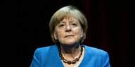 Angela Merkel, die ehemalige Bundeskanzlerin. Sie ist eine alte Frau mit etwas längeren Haaren, die ihr Gesicht umrahmen. Merkel trägt eine Kette aus großen, braunen, leicht transparenten Steinen oder Perlen.