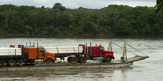 Eine Fähre auf einem Fluss im Regenwald mit LKWs, die mit Rohren beladen sind.