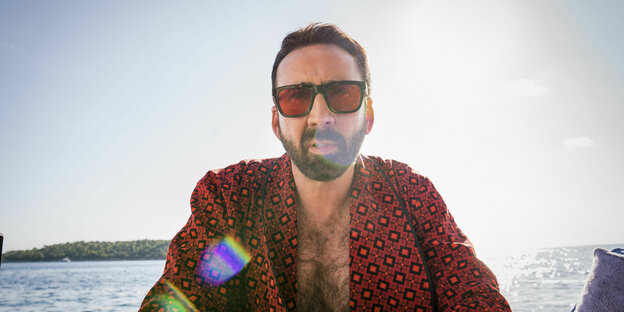 Nicolas Cage mit Sonnenbrille und im Bademantel mit Brustbehaarung vor dem Meereshorizont