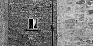 In einer Mauer aus Ziegelsteinen sieht man ein einzelnes Fenster, eine Frau schaut heraus