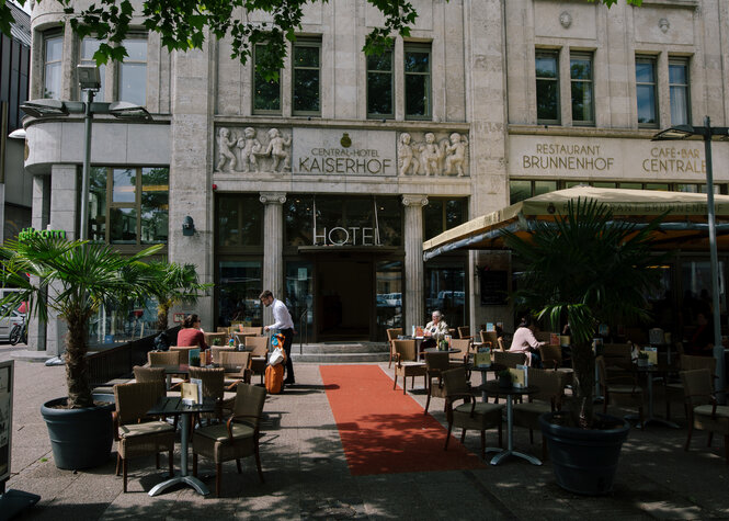 Ein schickes Hotel von außen, ein roter Teppich führt zum Eingang; draußen stehen Tische und Stühle, teilweise unter Schirmen. Ein Mann im weißen Hemd serviert an einem Tisch