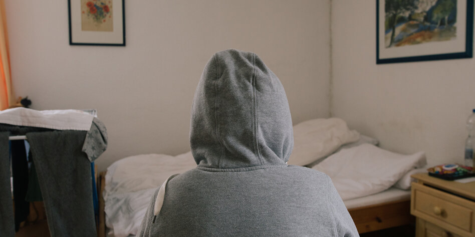 Eine Person in grauem Pullover mit übergezogener Kapuze sitzt mit dem Rücken zur Kamera. Dahinter sieht man den Raum: Ein Doppelbett und eine schimmlige Wand