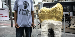 Ein junger Mann steht neben dem Model eines Goldzahns- der Slogan auf seinem T-Shirt: Lumumba still alive