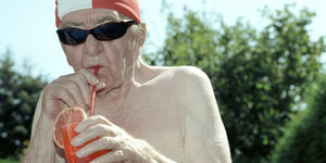 Ein älterer Mensch mit nacktem Oberkörper und Sonnenbrille saugt ein rotes Getränk aus einem Glas