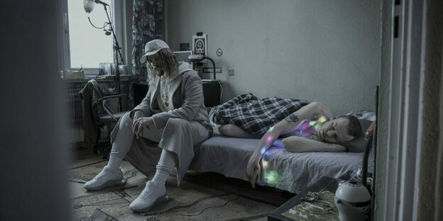 Szene aus „Other People“ (R: Aleksandra Terpińska, 2021): ein Mann sitzt in einer Plattenbauwohnung auf einem Bett. Daneben schläft ein anderer Mann.