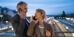 Ein Mann und eine Frau stehen dicht beieinander auf einer Brücke, sie dreht sich zu ihm um, sie schauen sich an, beide haben ein Handy am Ohr und scheinen zu telefonieren.
