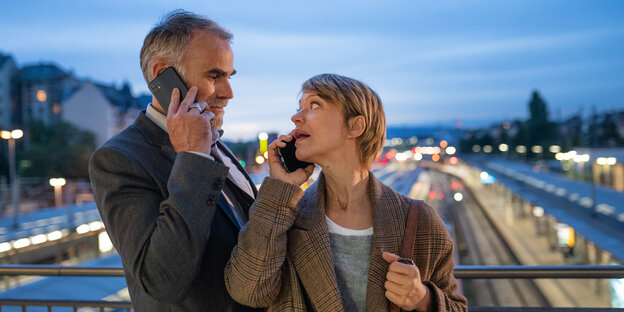 Ein Mann und eine Frau stehen dicht beieinander auf einer Brücke, sie dreht sich zu ihm um, sie schauen sich an, beide haben ein Handy am Ohr und scheinen zu telefonieren.