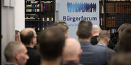Hinterköpfe von Männern bei einer Veranstaltung. In Blickrichtung steht vor einer Bücherwand ein Aufsteller mit der Aufschrift Bürgerforum