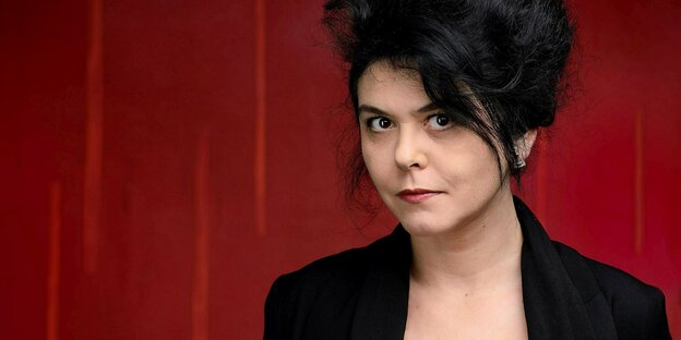 Die Autorin Chloé Delaume steht mit ihren dunklen Haaren zu einer Hochsteckfrisur gesteckt vor einer roten Wand und blickt in die Kamera