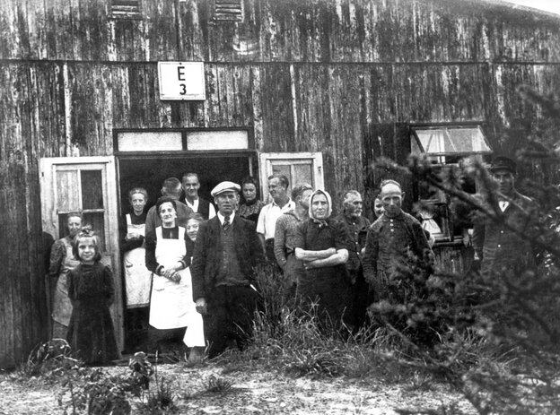 Ein schwarzweiß-Bild zeigt Menschen (Männer, Frauen und Kinder) vor einer Holzbaracke