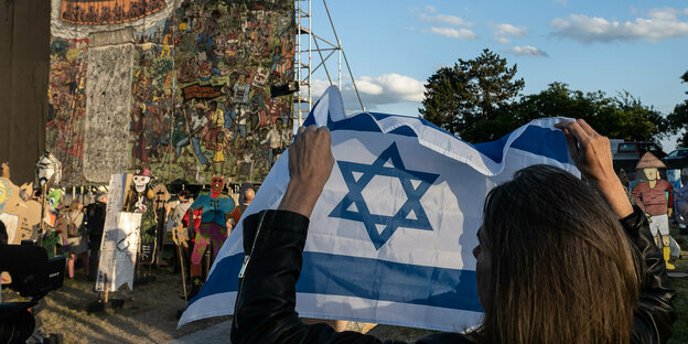 Eine Frau mit einer Israel-Flagge steht vor dem teilweise verhüllten, großflächigen Banner, eine Figurendarstellung "People·s Justice" (2002) des Kollektivs Taring Padi