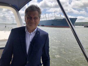Ein Mann mit kurzen grauen Haaren schaut in die Kamera und lächelt. Er trägt ein blaues Jacket und ein weißes Hemd. Er steht auf einem kleinen Boot. Hinter ihm ist ein großes anderes Schiff zu sehen. Das ist die Independence, das Gas-Terminal im Hafen von Klaipeda Ein Mann mit kurzen grauen Haaren schaut in die Kamera und lächelt. Er trägt ein blaues Jacket und ein weißes Hemd. Er steht auf einem kleinen Boot. Hinter ihm ist ein großes anderes Schiff zu sehen. Das ist die Independence, das Gas-Terminal im Hafen von Klaipeda