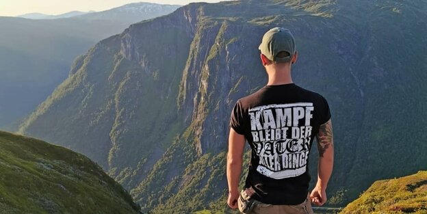 Ein Mensch steht vor einer Gebirgskulisse. Er trägt ein T-Shirt mit dem Text 