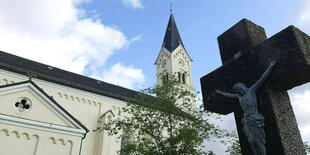 Eine weiße Kirche, davor ein schwarzes Kreuz mit einer Jesusfigur daran.