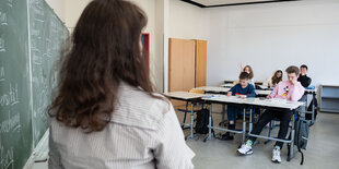 Eine Lehrerin steht an einer Tafel und blickt auf die Schüler hinter ihren Tischen.