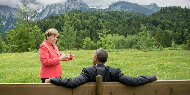 Angela Merkel steht vor Barack Obama der auf einer Bank sitzt.