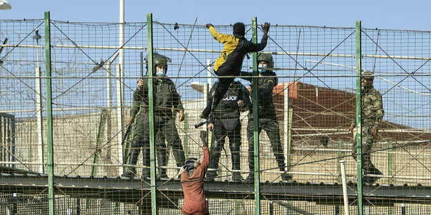 Zwei Männer versuchen über einen Grenzzaun zu klettern, der von Polizisten umzingelt ist