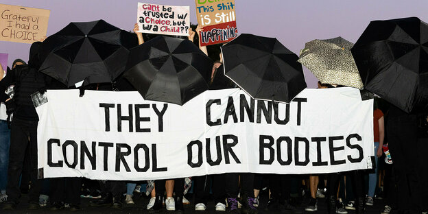 Menschen halten ein Banner mit der Aufschrift "They cannot control our bodies". Um das Banner herum sind sie durch schwarze Regenschirme verdeckt