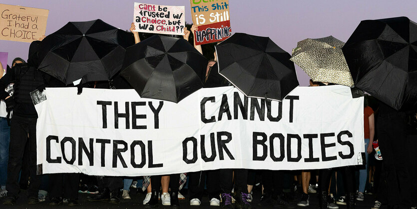 Menschen halten ein Banner mit der Aufschrift "They cannot control our bodies". Um das Banner herum sind sie durch schwarze Regenschirme verdeckt