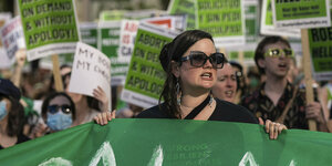 Eine Frau mit Sonnenbrille hält ein grünes Banner, im Hintergrund viele andere Protestierende mit Schildern